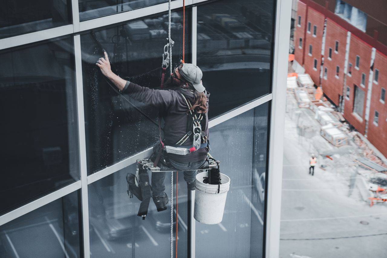 Un travailleur nettoie les vitres d’un gratte-ciel, équipé d’un harnais, dans un environnement urbain.